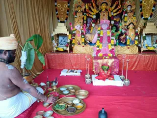 A seated priest with a bare upper body, white dhoti and a pale saffron turban sits at left on a red cloth, with white sacred ash marks on his chest and arms, performing a puja. Before him are brass plates and bowls holding flower petals, turmeric, kumkum, rice, betel leaves and other offerings, and a small brass pot with bamboo sticks. Behind him, a banana plant stands beside an image of a seated man in a framed black-and-white photo. The altar holds clay-like deity murtis of Durga with multiple arms, Ganesha and other figures, decorated with marigold and rose garlands. At centre a pink-robed figure with a pink turban stands on a pink-draped stool, also garlanded, while small framed portraits flank the altar and a red-bordered cloth covers the offerings in front.