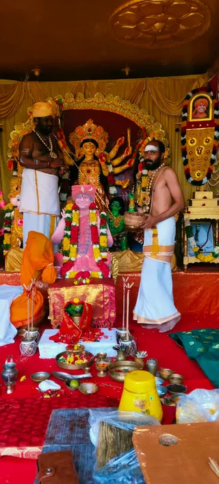 Photograph of a Navaratri Durga puja set up in a decorated shrine tent with gold drapes and a gilded arch. A multi-armed Durga murti stands behind a red-and-gold backdrop. In front, a large framed-style portrait printed on a pink cloth sits on a gold-brocade-covered platform, garlanded with marigolds and red roses. Two men stand at the shrine: on the left, a man with a yellow-orange turban, bare-chested, wearing a white dhoti and a beaded mala; on the right, a bare-chested man with a white dhoti and a white tilak, holding a brass vessel. A seated figure in orange robes sits at left near the platform. Red floor cloths hold brass plates, a kalasha-like vessel, flowers, fruit, lamps and offerings. A yellow container sits at lower right. A small framed photo garlanded with flowers sits on a side stand at right, and a gold-framed portrait hangs at upper right.