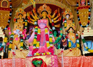 A photograph of a Durga Puja pandal shrine with a large multi-armed goddess murti in gold and red ornamentation, flanked by smaller painted deity figures including Ganesha on the left and a green-faced figure on the right. Foreground at centre, a framed photograph of a smiling man with a pink-and-red wrap and rudraksha mala has been placed over the central figure, garlanded with yellow, red and white flower garlands; the printed image is a depiction, not a physical person. Small framed portraits appear in the upper corners, one showing a man in an orange turban, and a red-flowered garland hangs at the bottom. Gold decorative panels, a red brocade altar cloth and offering items in front complete the scene.