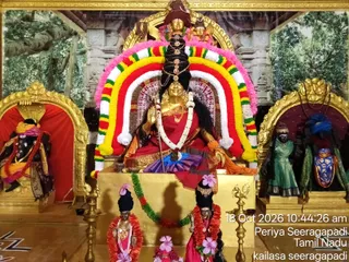 A photograph inside a temple shrine showing a golden deity murti of a goddess seated on a raised golden throne, dressed in red and purple garments with gold ornaments and a tall black hair crown. She holds a trident-like staff and a hand raised in blessing, framed by a large arch of red, yellow, green and white garlands. Flanking shrines hold smaller murtis: a dark figure in red and purple on the left, and a green-robed and blue-ornamented figure on the right. Brass oil lamps stand on the floor, with pink-flowered smaller murtis and garlands in front of the golden throne, and a painted forest backdrop behind. A timestamp reads 18 Oct 2026 10:44:26 am.