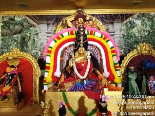 Photograph of a golden deity murti seated in a shrine at Periya Seeragapadi, Tamil Nadu, decorated with an arched garland of red, yellow, green and white flowers with pink edging. The central figure wears a red and gold sari, holds a trident and wears a long beaded necklace, with a tall dark hair crown above. Flanking niches hold a dark deity figure in red and orange on the left and a green-robed figure on the right. A backdrop shows painted forest and temple imagery, and oil lamps and floral offerings sit at the base. No physical people are visible.