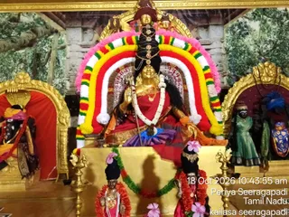 A decorated shrine photograph showing a golden deity murti of Mahadevi Kailasa Seeragapadi seated in a red and orange sari with a large raised arch of garlands in pink, yellow, red, green and white behind her. She holds a trident and raises one hand in blessing, wearing a gold necklace and a tall dark braided hairstyle with a crown-like ornament. Flanking deity figures stand in gilded niches, including a figure in a green garment on the right and a figure in red and gold on the left. Two smaller standing figurines in front wear flower garlands and pink blossoms, with brass oil lamps on either side and a green and red garland draped across the base. The backdrop shows painted temple architecture and foliage. No living people are visible.