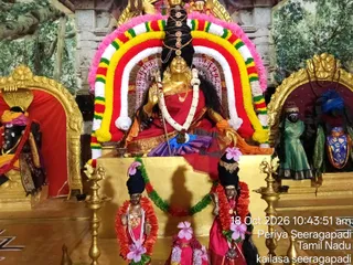 A photograph of a shrine with a seated golden-faced deity murti in a red and purple sari, one hand raised in abhaya mudra and holding a long staff, framed by a rainbow-coloured floral arch of yellow, red, white and green garlands with pink petals. Smaller standing murtis in front wear red and pink garments with pink flower garlands, and ornate brass oil lamps stand on either side. Painted forest backdrop, gold-framed niches holding dark-faced figures in green and blue garments, and a date stamp reads 18 Oct 2026 Periya Seeragapadi Tamil Nadu. No physical people are visible.