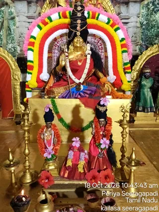 A decorated shrine in a temple hall showing a seated golden deity murti, a goddess figure with a tall black and gold crown and a trident held in one hand, wearing a red sari and garlands. Her arch behind her is framed with red, yellow, white and green garlands and pink flowers. In front stand smaller murtis of a male and female figure with black crowns, pink hibiscus flowers and orange and green garlands, along with a pair of red-clad figures. Brass oil lamps stand on the floor, one with a lit flame, alongside a brass pot and red hibiscus blossoms. Timestamp reads 18 Oct 2026 10:43:43 am.
