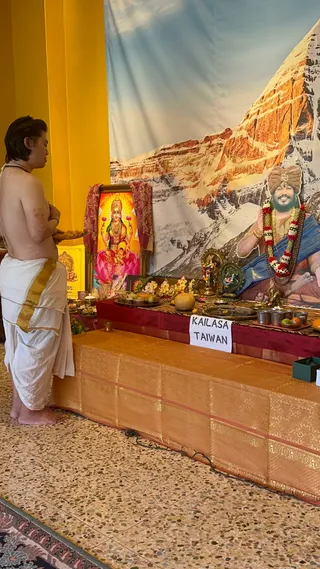 A bare-chested priest in white dhoti with a gold border stands at left, hands together in prayer before a shrine on a low table draped in gold and maroon fabric. Behind the table a large backdrop shows snowy mountain peaks, and a framed picture of a goddess seated on a pink lotus, with a red and gold paisley shawl, sits at center. On the right a large printed portrait of a bearded figure in a turban and blue shawl, with a garland of red and white flowers and rudraksha beads, is shown with a raised hand in blessing. A white card reads KAILASA TAIWAN. Brass lamps, fruit, a coconut, steel cups, small deity figurines and flower garlands are arranged on the altar.