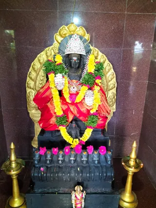 A black stone deity murti seated on a dark carved base decorated with pink flowers and small red stones, framed by an ornate gold arch. The figure wears a silver crown, red and gold silk garment, a red tilak, a large jewelled necklace, and garlands of yellow marigolds and green leaves with pink flowers. Two small silver-embossed hand pieces rest on the chest area. Two tall brass oil lamps with lit flames stand on either side, and a small brass figurine appears at the base. No people are visible.