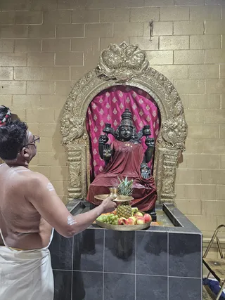 A bare-chested priest in a white dhoti with white sacred markings on his shoulders and forehead stands at left, offering a brass tray with a small brass cup to a black stone deity murti of Lakshmi seated in a golden arched shrine. The deity wears a deep red-maroon silk cloth and holds two pots or vessels in raised hands. Behind it hangs a magenta cloth with gold motifs inside an ornate gold-leaf arch. Before the murti on a black tiled platform sit a pineapple, green grapes, apples and other fruit on brass plates, with a small pool of liquid. The walls are gold-coloured block tiles and a folding chair is at right.