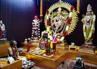 Inside a temple shrine with dark maroon drapes and a gold-toned platform, a priest with a shaved head and white body markings sits at left in a white dhoti and orange-brown shawl, holding a small silver vessel toward a gilded altar. At the centre, a golden arched prabhavali frames a deity murti adorned with a crown, silver ornaments and a long red garland. Flanking figures include standing murtis in a red turban and a crown with garlands, and a murti in green and gold with a silver necklace and white flower garland. Marigold and red garlands hang from above, with lotus flowers, brass plates and a kalasha placed around the altar, which suggests a puja in progress.
