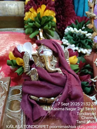 Close photo of a small brass Ganesha murti seated amid flower decorations. The figure wears a maroon cloth draped over its head and body, with a white tilak and red bindi on its forehead. Its raised hand holds a small object, and its trunk curls toward the lower right. Behind it are garlands of red, yellow and green paper-style flowers, white jasmine strings and a red-and-white woven fabric. A carved wooden surface is visible at the lower left. Printed text across the lower right corner gives a date stamp and address. No people are visible.