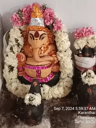 Photograph of a shrine niche holding a terracotta-coloured Ganesha murti seated at the centre, with a tall gold conical crown, silver foil band, trunk resting toward a bowl, red tilak on the forehead, and pink and magenta fabric trim at the waist. A garland of white jasmine-style flowers frames the figure, and pink flowers sit atop the crown. Two dark shivalingam-type stones with white flower garlands and red-marked tops stand in front and to the right, one draped in white cloth. Timestamp overlay reads Sep 7, 2024 5:59:37 AM Karanthai Thanjavur Tamil Nadu. No people are visible.