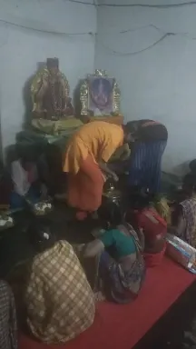 A photograph of a small indoor shrine room with white walls and a red floor covering. On the left a gold-coloured deity murti stands on an altar, and on the right a framed picture of a deity with a blue face sits in a decorated gold frame. In the centre a person in an orange-saffron top and dark pants bends forward over a pot or vessel on the floor. Near them a person in a blue garment kneels, and in the lower foreground people in checked cloth and red and teal saris sit on the floor among brass vessels and offerings, consistent with a food offering.