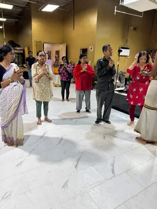 Photograph inside a spacious hall with yellow walls and white marble floor, where several devotees stand with palms pressed together in namaskar. A woman in a red floral kurta holds a small brass vessel and raises her hand to her head, appearing to receive blessings from a priest in white dhoti with a gold border at the right edge. A woman in a lavender-bordered white saree and a woman in a green floral tunic stand at left, and a man in a black striped shirt and grey trousers stands center-right. Behind them a robed figure in white stands near a shrine area with deity murtis and altar items. Visible people are roughly ten, including partially visible figures in the background.