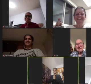 A five-panel video call grid of smiling participants in home settings. Top left, a woman with short grey hair, sunglasses and a dark red top, labelled VERA LUCIA, BRASIL. Top right, a woman with short grey hair and a red bindi in a white top beside a small figure on a shelf, labelled YOGAMALARI. Middle left, a woman with dark hair and a white sweatshirt with printed text, labelled Ma aradhana. Middle right, a smiling older man in a checked shirt, labelled Bob. Bottom centre, a woman in black with hands joined in namaste in front of a painting and a garland-draped frame, with a partly visible name label. Screen-captured participants are not physical people in the room.