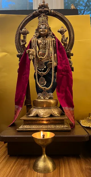 A photograph of a bronze deity murti of Venkateshwara standing before a brass arch-shaped halo, with four raised hands and a tall jeweled crown. The murti wears layered pearl and red-stone necklaces, a red-and-gold draped cloth hanging on both sides, and a dark garment. It stands on a lotus-petal base on a wooden pedestal against a golden backdrop. In the foreground a brass oil lamp with a lit flame burns on a wooden floor. No people are visible.