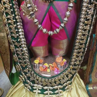 Close view of a deity murti dressed in a magenta silk garment with gold zari border and a green velvet cape embroidered with silver-gold floral motifs and round silver studs. A heavy silver necklace of bead-like links hangs across the chest. At the base, a silver pedestal holds a cluster of orange, yellow and pink flowers arranged around silver cups, with a gold-brocaded fabric draped below.