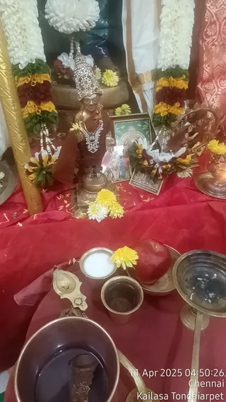 A photograph of a ritual altar draped in red cloth, with a small metal deity murti at its centre wearing a silver crown and a silver necklace, dressed in a red garment and holding a yellow flower. Yellow and white flowers, white and red garlands with yellow and green accents hang on the gold-coloured pillar and white cloth backdrop. A framed picture stands behind the murti. In the foreground are a brass oil lamp, a small bowl of white liquid, a brass cup of water, a red apple, a brass pot of water and a tall brass lamp stand. This appears to be a puja or daily ritual setup with offerings. A timestamp in the corner reads 11 Apr 2025 04:50:26, with the labels Chennai and Kailasa Tondaiarpet.