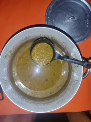 Overhead photo of a large aluminium cooking pot filled with a yellowish lentil or split-pea stew, with a black ladle lifting a portion of the thick soup. The pot's lid rests against its rim at the upper right, set on a bright orange tablecloth. No people or a person visible in the frame; only a hand edge appears at the right.