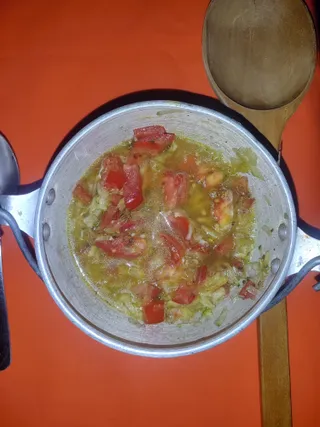 Overhead photo of a silver aluminum pot on a bright orange surface containing a cooking mixture of chopped tomatoes, diced green vegetables and onion in a yellow oily broth with seeds visible. A large wooden spoon rests at the upper right and a metal spoon handle appears at the left edge. No people are visible.