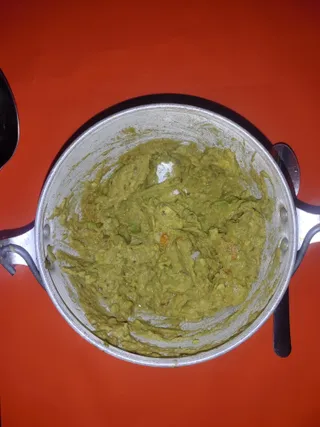 Overhead photo of a silver aluminium pot filled with a smooth, mashed green paste. The pot has two looped side handles and sits on a bright red surface. A dark spoon rests at the right edge and part of another dark object appears at the upper left. No people are visible.