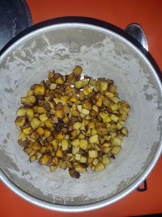 A top-down photo of a large stainless steel pot on an orange surface, filled with diced fried potatoes that are golden yellow and browned at the edges. A metal ladle rests on the rim at upper right, and a dark pan lid sits at the upper left. No people or deity images are visible; the scene shows only the food in the vessel.