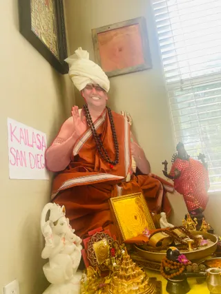 A seated figure in an orange-red saffron robe with cream-bordered edges and a white turban sits in a corner beside a window with white blinds, one hand raised in blessing. A dark rudraksha mala hangs on the chest with a small red tassel. Before the figure stands a white marble Ganesha statue on the left, a golden framed yantra plate in the centre, and a brass tray of rudraksha malas, pink flowers and a small black lingam with a metal pot in front. A golden pyramid-shaped shrine piece and a red-dressed dark statue stand to the right. A handwritten pink paper sign reading KAILASA SAN DIEGO hangs on the left wall. Deity murti and altar objects surround the figure.