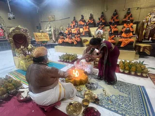 A photograph inside the KAILASA Toronto temple hall during a fire ritual. A bare-chested priest with a saffron head wrap and gold-bordered white dhoti sits on a low red stool at the left, tending a flame over a yellow-and-gold homa vessel. Beside him, a seated woman in a white sari and a second woman in a magenta sari bend forward over the fire, one offering something to the flame. Brass plates, bowls, and a small kalasha lie on the blue-and-white rug in front, with rose petals and banana-leaf offerings scattered around. Behind them, a row of black deity murtis in saffron robes sits on tiered golden platforms against a plain wall. A white sign reads KAILASA TORONTO CANADA. A gilded throne with a red curtain stands at left, and a deity murti draped in flower garlands stands at right. No a person candidate is visibly present.