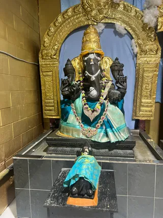 A photograph of a shrine niche with a large black stone murti of Ganesha, seated with four arms, adorned with a gold crown, jeweled necklaces, and a turquoise silk cloth draped over the seat. A gilded arched frame with floral carvings surrounds the deity, and a blue backdrop with cotton-like decorations stands behind. In front, on a black stone platform, a smaller black figure, possibly a devotee or attendant statue, is draped in a teal and yellow cloth. No living people are visible.