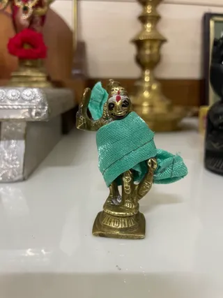 A small brass deity murti stands on a square base on a white surface, draped in a teal-green cloth wrapped over its shoulders and arms. The figure has a red tilak mark on its forehead and raises one hand in a gesture, with the other arm bent at its side. Behind it are blurred brass lamp stands, a red flower in a vase at left, and a dark object at right. No people are visible.