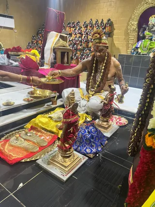 A seated man in a tall gold-and-red ornate headdress, bare-chested with a white dhoti edged in gold, wears layered rudraksha malas and a heavy gold necklace with red tilak on his forehead. He holds a brass lamp-like vessel in one hand while a second person's arm extends from the left, offering a plate with a lit lamp. Before him on a black tiled floor stand two gold-finished deity figures, one in red and one in maroon, with a yellow and blue cloth and brass ritual items laid out. A wall of small black murtis in orange and white garments, floral garlands and a green-clad deity image complete the shrine setting.