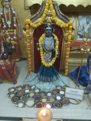 A photograph of a shrine altar with a dark blue-skinned deity murti wearing a teal silk skirt, silver-white beaded necklace and jewelled waist ornaments, garlanded with yellow and orange marigolds and set within a gold arch. Around its base sit about thirty small steel bowls filled with grains, pulses and seeds arranged in a circle. A brass trident stands at right beside a white sign reading KAILASA THIRUCHENGODU and ERODE. On the left, a standing life-size cutout of a smiling figure in orange robes and rudraksha garlands, and a seated dark statue with a garland. A framed black-and-white portrait of a bearded sage hangs in the upper right. A small oil lamp burns in the foreground. Timestamp overlay reads 03-Oct-2024 05:41:22.