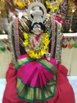 A seated deity murti. It wears a pink and green silk sari with gold zari borders and a jewelled ornamental crown with a halo-like backdrop. Yellow flower garlands drape its shoulders and chest, with gold necklaces and a long green stalk held in one hand. Behind it, a brass oil lamp and gold ceremonial items stand against a patterned backdrop, and red fabric covers the seat.