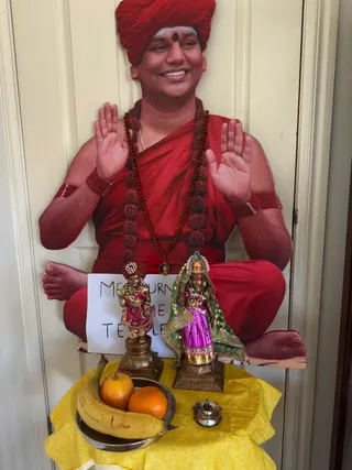 A life-size cutout of a smiling man in a red turban, red robe and red wrist bands, seated cross-legged with both palms raised, is displayed on a cream wooden door. A dark bindi and a long rudraksha mala with a small pendant hang on his chest. In front stand two brass deity murtis on a yellow cloth-covered table: the left one in a red turban with a pink dress, the right one in a green veil and pink skirt. A stainless steel plate holds a banana, an apple and an orange, and a small brass oil lamp sits beside it. A handwritten paper sign reads Melbourne Temple.