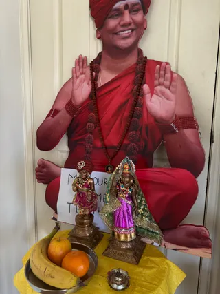 A small household shrine on a yellow cloth in front of a cream door. A large cutout photo of a smiling man in a red turban, red robe and long rudraksha mala sits in a lotus pose with palms raised, labelled as a depiction on a printed card. Two bronze deity figures stand in front: a small one in a red turban and pink garment, and a larger female figure in a purple sari with a green embroidered veil and jewelled crown. A silver plate holds a banana, an apple and an orange, and a small brass oil lamp sits in front. No physical people are present.