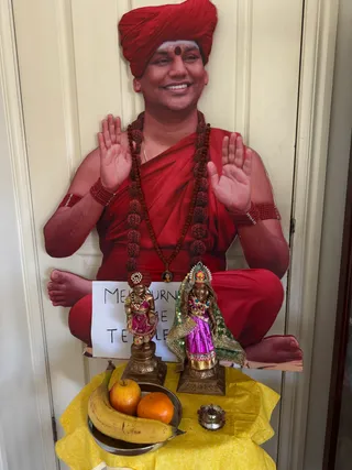 A photograph of a home shrine on a cream-painted door. Centre back is a large life-size standing cutout of a smiling man seated cross-legged in red, wearing a red turban with a red bindi, a red shawl, a long rudraksha mala and red beaded bracelets, with both palms raised in blessing. Printed on the cutout's torso is a small portrait pendant. In front, a white paper sign reads Melbourne Home Temple. Two small bronze deity murtis stand on a yellow cloth: the left one is a child-form figure in a red turban and pink garment, the right one a female form in a green-and-gold veil, pink skirt and a jewelled headdress, with beaded necklaces. Beside them are a steel bowl of fruit (a banana, an apple and an orange) and a small silver oil lamp. The image is of the cutout and statues only, with no physical people present.