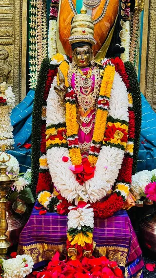 A golden metal murti of a goddess, presumably Devi Rajarajeshwari as named in the page excerpt, stands centrally, wearing a tall golden crown and a red-pink garment with ornate jewelled necklaces. She is draped in dense garlands of white, yellow-orange and red flowers, with green leaf garlands framing her. She holds a lotus-like emblem in one raised hand. She sits on a purple and gold brocade cloth, surrounded by scattered red and pink petals, white flower bunches, and a brass oil lamp on the left. Behind her hang an orange and gold fabric, a teal silk drape, and ornate gold-toned panels. No people are visible.
