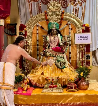 A shirtless priest in white dhoti with orange-gold border and sacred marks on his bare back and arms stands at left, reaching toward a seated golden-bronze deity murti of a goddess. The murti wears a green turban-style crown, a red and white jeweled necklace, a gold and green sari with pink cushion behind, and white and red garlands. In front stand a brass kalasha-style vessel with flower garlands, a red decorated pot and a copper pot with yellow flowers. A gold ornate arch frames the shrine, with a placard reading KAILASA SAN JOSE on a stand to the right. The scene depicts puja at a shrine.