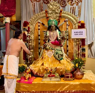 A priest with a bare upper body and a white dhoti bordered in gold stands at left, holding a lit oil lamp toward a seated deity murti. The murti wears a green turban, a gold and red-jewelled necklace, a magenta cloth and a yellow-gold skirt with green trim, and is framed by a gold arched throne with a garland of red and white flowers. In front are brass lamps, a flower-decorated vessel, a copper pot and a yellow floral arrangement on a gold cloth. A framed sign reads KAILASA SAN JOSE beside the throne. The scene shows a puja.