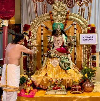 A bare-chested priest in a white dhoti with a gold border stands at left, lit oil lamps in his hands, performing aarti before a seated deity murti of Devi Rajarajeshwari. The murti wears a green turban, a gold and rose-coloured sari with green border, layered jewelled necklaces and a red cushion beneath her. She sits on an ornate gold peetha framed by an arched gold torana. Marigold and rose garlands hang around the throne, with a brass kalasha offering, flowers in a copper pot, and a sign reading KAILASA SAN JOSE beside the shrine. No physical a person candidate is visible.
