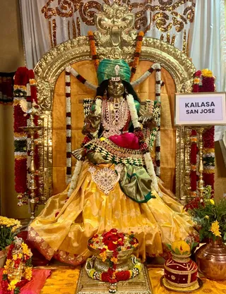 A seated metallic murti of Devi Rajarajeshwari is the central subject, dressed in a golden-yellow silk sari with a green and gold border, ornate jewelled necklaces, a red cloth at the lap and a green turban-style crown. Garlands of marigold, white jasmine and red flowers drape the figure and the golden arched peetha behind it. In front, a brass vessel holds red rose petals and flowers, a red and gold kalasha sits to the right, and a copper pot and yellow chrysanthemums fill the foreground. Brass oil lamps flank the throne. A printed white sign reading KAILASA SAN JOSE stands at the right edge. No people are visible.