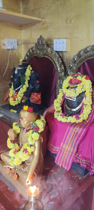 A small shrine corner photographed indoors against a yellow-ochre wall with a wall switchboard above. On the left a black stone Ganesha murti sits on a red cloth, garlanded with yellow marigolds and wearing a red cloth. On the right a black Shiva lingam stands on a magenta cloth, wrapped in yellow marigold and red rose garlands with a gold-coloured band and a rudraksha-style bead arrangement. In the foreground a golden seated statue of a figure with hands pressed together in anjali, wearing a turban, is draped in yellow and orange marigold garlands with red roses. A small brass oil lamp with a lit flame sits at the bottom left. No people are visible; the scene shows puja decoration of the murtis.