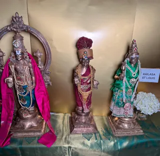 Three metal murti statues stand on a gold-toned draped cloth with green and gold brocade on a table. The left figure, with a crown and pearl and gem necklaces, wears a magenta shawl and a teal skirt, and holds its hands in a blessing pose. The central figure wears a maroon turban with jewelled ornaments, gold necklaces and a maroon dhoti. The right figure wears a green blouse and teal skirt with pearl necklaces and a jewelled crown, holding a staff. A white hydrangea-like flower arrangement sits at the right, and a small white card reads KAILASA ST LOUIS. A gilded backdrop with an arched frame stands behind them.