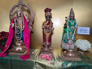 Three bronze deity murti statues stand on a green and gold brocade cloth before a gold backdrop. The left figure, under an arched frame, wears a pink shawl, blue skirt and long pearl and bead garlands. The centre figure wears a maroon turban and maroon dhoti with a gold-and-jewelled necklace and garlands, one hand raised in blessing. The right figure wears a green blouse and skirt with pearl and red-stone necklaces and holds a kalasha-like vessel. Red chrysanthemums, a white hydrangea arrangement and scattered pink petals sit at their bases. A white placard reads KAILASA ST LOUIS.