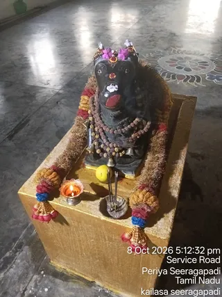 A photograph of a black stone Nandi bull murti seated on a gold-painted pedestal inside a hall with a polished grey floor. The murti wears a rudraksha mala with brass bells, a pink and purple flower crown, a red tilak and white flower on its face, and fringed garlands in red, gold and blue draped over the pedestal. A small lit oil lamp in a silver dish sits on the left, with incense sticks in an ash bowl and a yellow fruit near the base. The lower right corner carries a timestamp and location overlay. No people are visible.