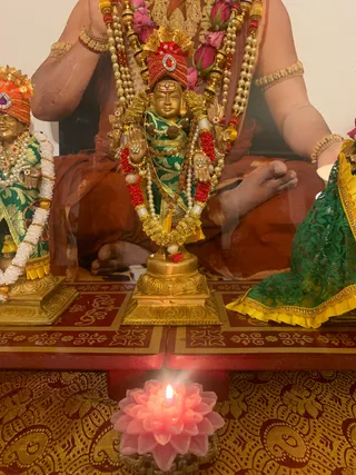 A photograph of a shrine altar with gilded metal deity murtis dressed in green and gold silk, adorned with red and white garlands, pearl strands, rudraksha beads and flower garlands with pink roses. A central standing murti wears a red and gold crown and a tilak on the forehead, with raised hands. Flanking murtis wear orange-red and gold crowns, and one is draped in green lace with a yellow border. Behind them a large painted or printed portrait shows a seated figure in orange-red robes, with bangles on the upper arm. A pink lotus-shaped candle with a lit flame sits in front on a red and gold brocade cloth. No people are visible in the scene.