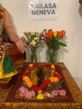 A photograph of a puja setup on a gold-and-maroon brocade cloth. Two wooden ritual objects with red and yellow marigold garlands sit on a maroon cloth with pink petals and a yellow flower. Behind them, two glass vases hold pink, yellow and orange roses and carnations. A printed paper sign on the wall reads KAILASA GENEVA. At the left edge, a seated person in an orange-brown garment wears a pearl and red bead necklace and gold bangles; a green and yellow silk garment is visible below. Only partial limbs are visible, so the person is not clearly identifiable.