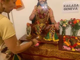 A woman in a pale yellow top stands at left, lighting an oil lamp or flame on a small altar. The altar has a red and gold brocade cloth and holds three gilded deity murtis dressed in green and yellow garments with white and red garlands; the centre figure wears a crown and a large gold necklace. Behind them is a large printed portrait of a bearded man in saffron-orange robes with a pearl and rose garland, a gold armband and a raised hand in blessing; he is shown on a screen or print, not physically present. To the right are vases of pink, orange and yellow roses and a small tray of marigold garlands and rose petals. A white sign reads KAILASA GENEVA on the wall.