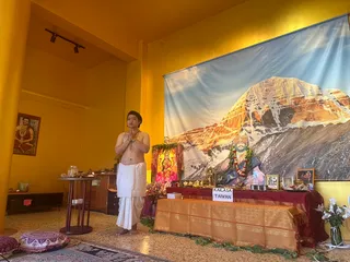 A photograph of a shrine room with yellow walls and a large backdrop of a snow-covered mountain peak in warm sunlight. A bare-chested man with short dark hair stands at the left in a white dhoti, palms pressed together in namaskar, with a rudraksha-style mala around his neck. On the altar draped in red and gold cloth, a sign reads "KAILASA TAIWAN"; there are flowers, brass deity figures, a framed portrait with garlands, and a small framed picture. A small framed image hangs on the left wall, and cushions lie on the floor.