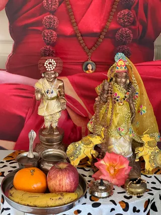 A home altar photograph showing two bronze-toned deity murtis in front of a large red fabric backdrop with a printed image of a seated figure in red robes. The figure on the left is a small standing murti with a maroon turban decorated with white pearls, wearing a cream and gold garment and holding one hand raised in blessing. The figure on the right wears a yellow embroidered sari with a veil and a red and gold headdress, carrying a small object and draped with rudraksha beads and a multicolored bead garland. Two yellow painted elephants flank the right murti. A rudraksha mala hangs at the top of the backdrop with a small round pendant bearing a photo of a smiling woman. In front, a silver plate holds an orange, an apple and a banana, with a pink rose, a silver oil lamp with a lit wick, a small silver pot and a silver cup with a spoon. The table is covered with a leopard-print cloth.
