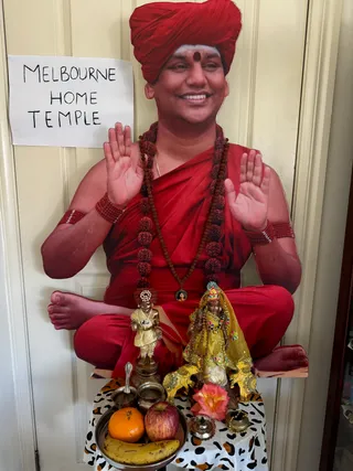 Photograph of a home temple altar set against a cream panelled door. A large cut-out life-size figure of a smiling man in a red turban, red robe, and multiple rudraksha malas and beaded red wrist bands sits cross-legged with palms raised, a red bindi and vibhuti on the forehead. A small paper sign reads Melbourne Home Temple. In front, on a leopard-print cloth, stand two gold-coloured metal deity murti dressed in yellow and green garments, one holding a flute, with an orange, an apple and a banana on a steel plate, a pink flower, a brass kalasha and an oil lamp. The figure cut-out is a printed image, not a physical person, so no people are present.