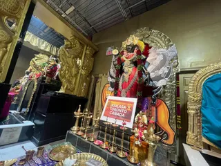 Interior shrine in a temple hall with a large dark-skinned deity murti standing on a black pedestal, adorned with red and green garlands, gold jewellery and a red-and-gold garment, holding a trident and other implements. A printed sign in front reads KAILASA TORONTO CANADA. Brass oil lamps with lit wicks, brass vessels and a tray with flower petals sit on the altar step. Ornate gold-coloured carved panels and a golden lion-form sculpture frame the shrine, and a colourful bouquet of flowers is at the base. No people are visible.